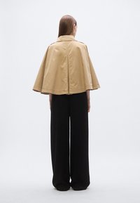 Bež kaput u stilu plašta s ovratnikom, s prorezom na leđima i širokim rukavima, uparena s crnim širokim hlačama. Glatki materijal, minimalistički dizajn.