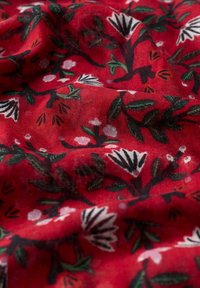 Sarkans audums ar ziedu rakstu, kurā redzami melni zari, zaļas lapas un baltas ziedi, parādot krokas un tekstūru.