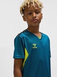 Dreng med korte blonde dreadlocks iført en teal og gul Hummel sportstrøje med kædehalskæde mod grå baggrund.