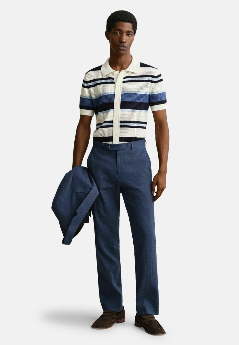 Uomo che indossa una polo a righe a maniche corte in maglia, pantaloni blu su misura, mocassini marroni, con in mano un blazer blu abbinato.
