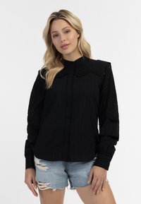 Zwarte blouse met gekraagde kraag en ge textureerde kanten mouwen, met een knoopsluiting aan de voorkant en een ribbelstructuur in het lichaam, gecombineerd met versleten denim shorts.