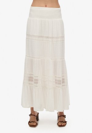 Witte gelaagde maxirok met kanten inzetstukken, gerimpelde tailleband, gedragen met bruine sandalen met hak, getoond van taille tot voeten op een effen achtergrond.