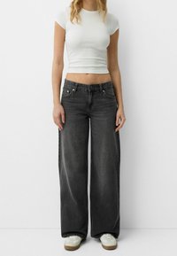 Černé džíny s širokými nohavicemi a vybledlým vzhledem, spárované s přiléhavým bílým tričkem. Součástí outfitu jsou světlé tenisky s texturovanou podrážkou.