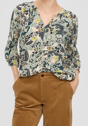 Woman wearing a multicolored abstract floral blouse with three-quarter sleeves and brown corduroy pants with hands in pockets.