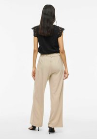 Donna con lunghi capelli scuri che indossa un top a maniche corte in pizzo nero, pantaloni beige a gamba larga e sandali neri con tacco alto, in piedi su uno sfondo bianco.