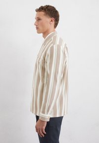 Gestreepte blouse in beige, witte en groene tinten; beschikt over een knoopsluiting bij de kraag, een borstzak en gedetailleerde manchetten. De stof lijkt getextureerd.