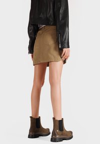 Stivali alla caviglia in suede marrone con pannelli elasticizzati ai lati, abbinati a una giacca di pelle nera e una gonna di jeans beige. Texture liscia, design casual.