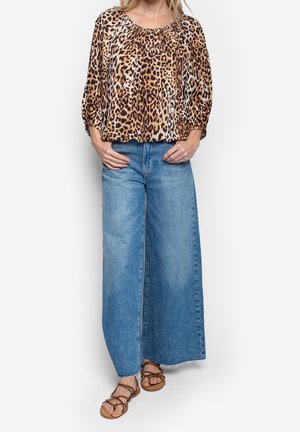 Femme portant un chemisier à imprimé léopard, un jean bleu à jambes larges et des sandales marron à brides, debout sur un fond uni.