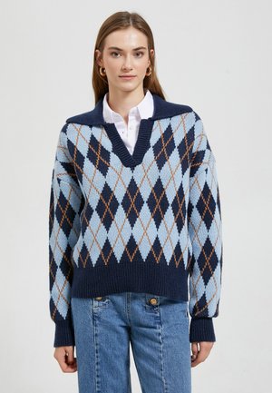 Sweter dzianinowy z niebiesko-granatowym wzorem w diamenty, dekoltem w serek i prążkowanymi mankietami. Połączony z jasnoniebieskimi dżinsami.