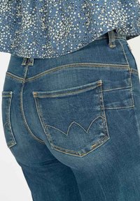 Jean en denim bleu foncé, avec une coupe ajustée, deux poches arrière avec un motif de couture unique en forme de feuille et des accents de couture en jaune doré.