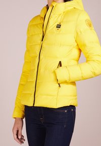 Giubbotto imbottito giallo con zip frontale, cappuccio e tasche laterali. Presenta un design trapuntato e una toppa con logo sulla spalla. Tessuto liscio e lucido.