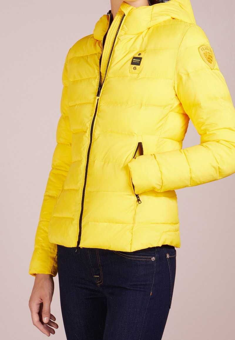 Giubbotto imbottito giallo con zip frontale, cappuccio e tasche laterali. Presenta un design trapuntato e una toppa con logo sulla spalla. Tessuto liscio e lucido.