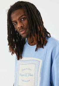 Camiseta azul cielo con un patrón de paisley en blanco y texto en azul. Manga corta, cuello redondo, hecha de una tela suave y ligera.