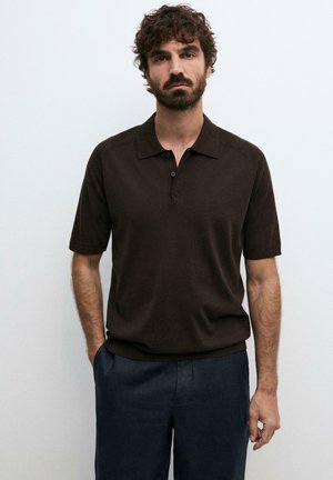 Uomo con capelli ricci e barba, indossa una polo marrone scuro a maniche corte e pantaloni blu navy, in piedi davanti a uno sfondo chiaro e uniforme.