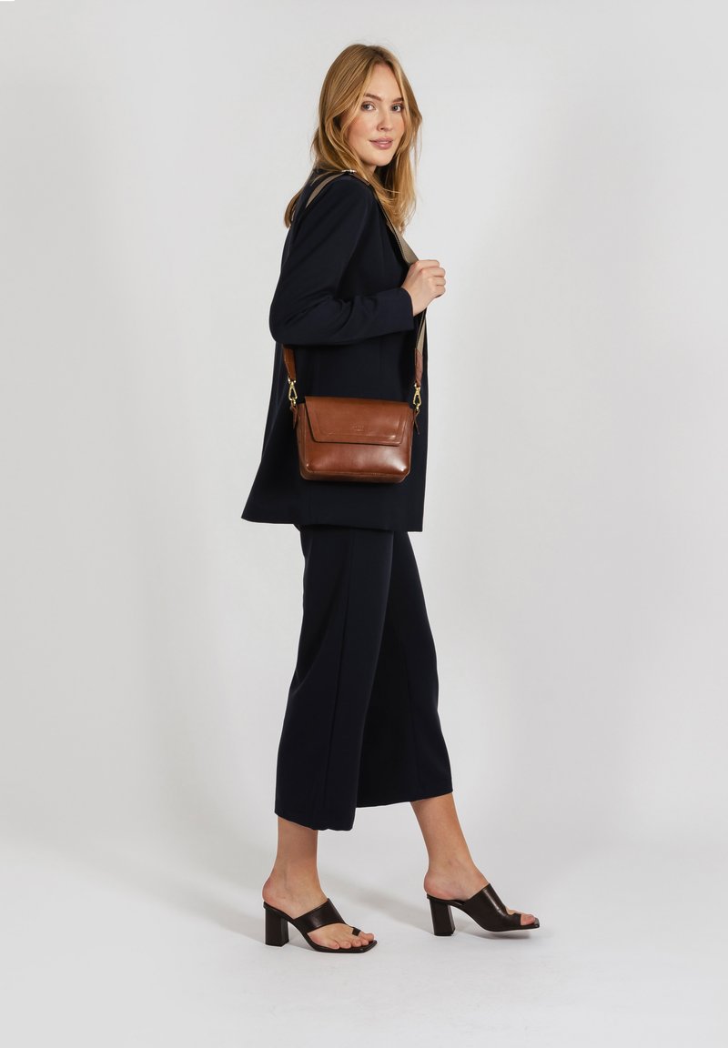 Borsa a tracolla in pelle marrone, dalla forma piatta, con chiusura zip e tracolla regolabile, indossata in modo casual su un outfit scuro.