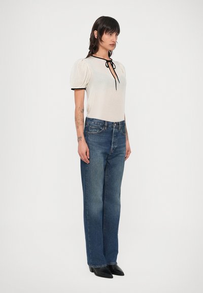 Λευκό υφασμάτινο τοπ με φουσκωτά μανίκια, μαύρο περίγραμμα και λεπτομέρεια δεσίματος. Φορεμένο με ψηλόμεσα μπλε τζιν και μαύρες μυτερές παπούτσια.