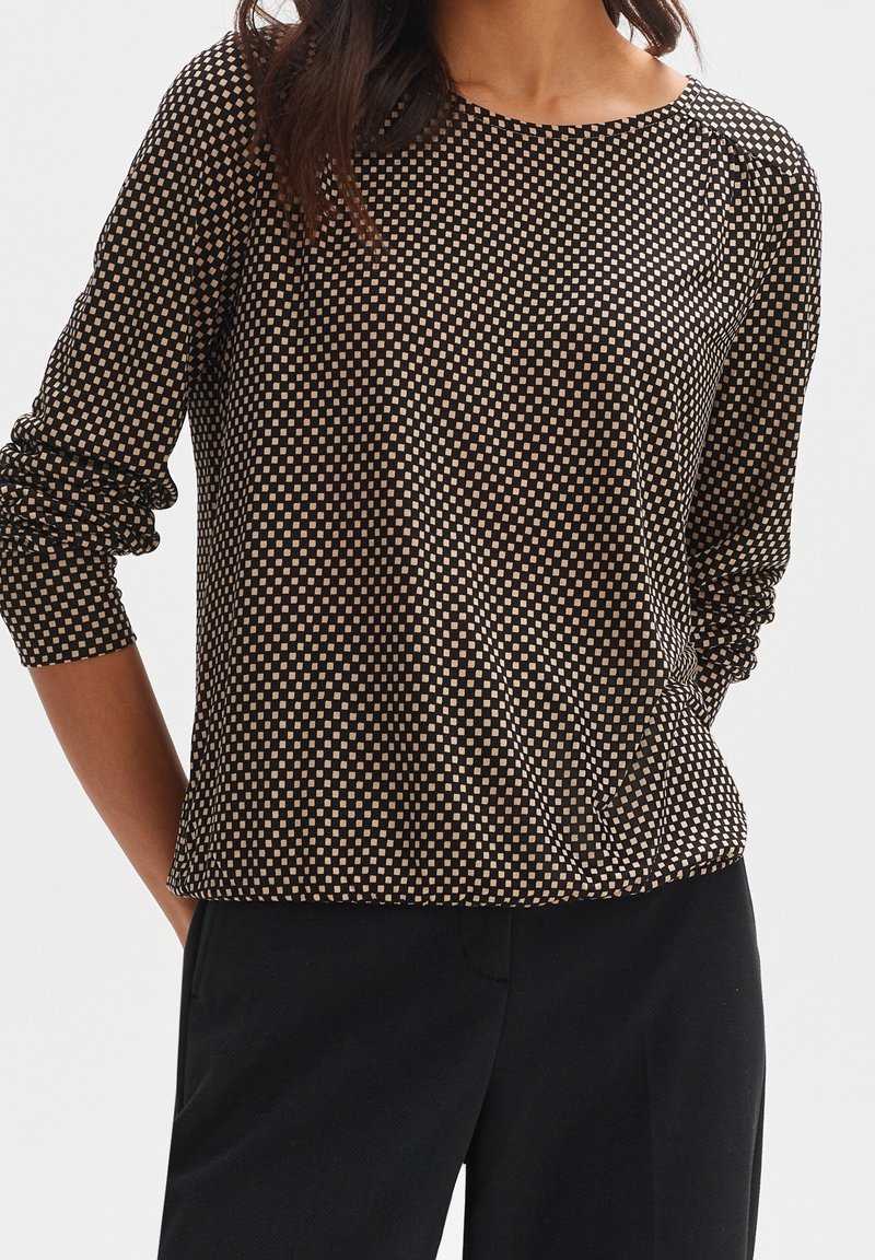 Femme portant un haut noir à manches longues avec un petit motif carré beige et un pantalon noir, debout les mains dans les poches.