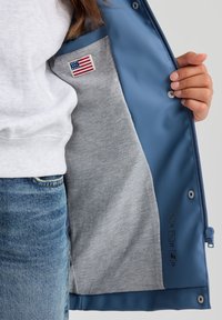 Giacca impermeabile blu con fodera in pile grigia. Presenta un patch della bandiera degli Stati Uniti, chiusura a zip e simboli di cura sul lato interno.