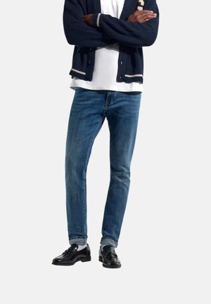 Persona che indossa un cardigan blu scuro, camicia bianca, jeans blu, scarpe eleganti nere lucide, in piedi con le braccia incrociate contro uno sfondo semplice.