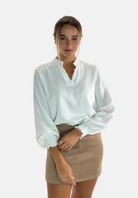 Weiße Bluse mit langen Ärmeln, Halbknopfleiste und gerafften Manschetten, kombiniert mit einem beigen Minirock, der eine glatte Textur zeigt.