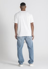 Camiseta blanca de algodón con mangas cortas, combinada con unos vaqueros holgados de color azul claro. Exhibida desde la parte trasera, mostrando un ajuste relajado y textura.