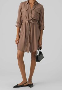 Abito button-down color beige realizzato in tessuto morbido, con cintura a nodo, due tasche sul petto e maniche arrotolate. Abbinato a ballerine nere con cinturino e una borsa.