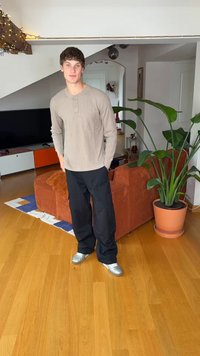 Jauns vīrietis tērpies gaiši brūnā, garroku Henlija kreklā un melnās, brīvāka piegriezuma biksēs ar sudraba krāsas apaviem stāv netālu no auga un dīvāna.