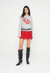 Γκρι πουλόβερ με κόκκινο γραφικό σχεδιασμό, φορεμένο πάνω από κόκκινη φούστα με πουά. Μαύρες μπότες μέχρι το γόνατο, με μυτερή μύτη και χαλαρή γραμμή.