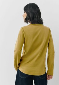 Langærmet top i sennepsguld, lavet af blødt stof. Har en rund halsudskæring og en afslappet pasform med sidesømme.