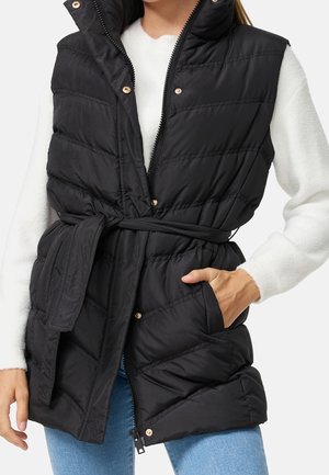 Femme portant une veste sans manches matelassée noire avec ceinture, un pull blanc à manches longues et un jean bleu, une main dans la poche de la veste.