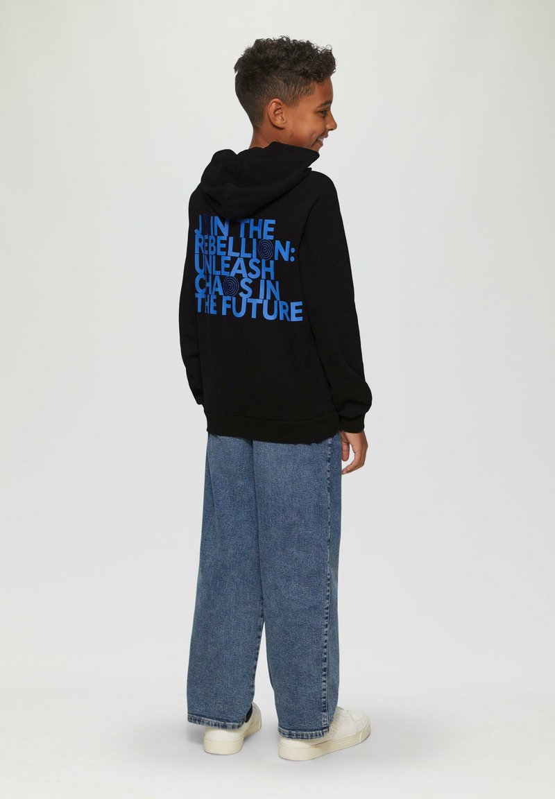 Zwarte hoodie met blauwe tekst op de achterkant waarin staat: "Sluit je aan bij de Rebellie: Laat Chaos in de Toekomst Vrij". Gecombineerd met lichtblauwe wijde jeans.