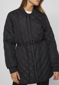 Vadderad svart jacka med dragsko i midjan, ribbade muddar och rund halsringning. Tyget är slätt med en blank yta.