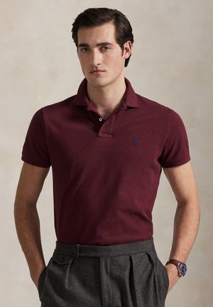 Jeune homme aux cheveux foncés portant un polo bordeaux et un pantalon plissé gris, mains dans les poches, fond neutre.