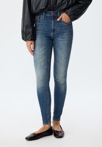 Mršave plave traperice srednjeg pranja, naglašene kopčanjem na dugme i prednjim džepovima. Nosive s tamnim ravnim cipelama koje imaju jednostavan detalj mašne.