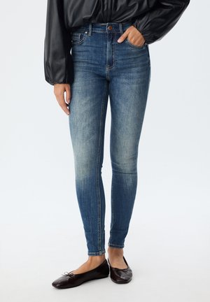 Tanke modre kavbojke z zmerno pranjem, okrašene z gumbom in sprednjimi žepi. V kombinaciji s temnimi ravnimi čevlji, ki imajo preprost detajl v obliki mašne.