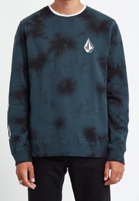 Sweatshirt crewneck en tie-dye vert-bleu avec des accents noirs, comportant un patch logo sur le devant et des poignets et un ourlet côtelés. Texture lisse.