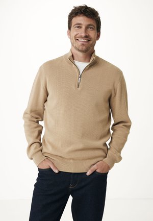 Maglione beige con zip in morbido materiale a maglia. Caratterizzato da un colletto alto, polsini e orlo a coste. Indossato con jeans scuri, mani nelle tasche.