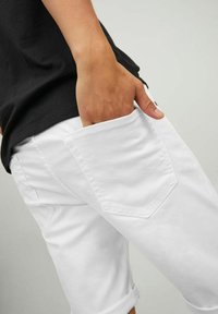 Shorts de algodón blancos con dobladillos enrollados, que presentan un bolsillo trasero con un sutil diseño cosido. Combinados con una camiseta negra.