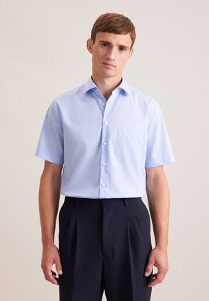 Jeune homme portant une chemise bleu clair à manches courtes avec une poche poitrine et un pantalon foncé à taille haute sur un fond uni.