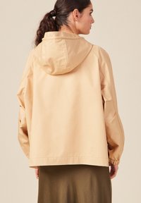 Giacca beige oversize con cappuccio, caratterizzata da una texture liscia e maniche elastiche, vista dal retro su uno sfondo neutro.