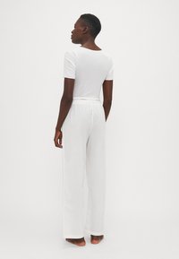 Weißes Baumwoll-T-Shirt mit kurzen Ärmeln, kombiniert mit lockeren weißen Hosen, die einen elastischen Bund mit Logo-Details aufweisen.