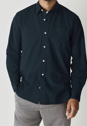 Homme portant une chemise bleu marine foncé à manches longues boutonnée avec une poche poitrine et un pantalon gris clair, debout, les mains détendues.