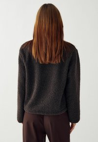 Persona con capelli castani lisci e lunghezza spalle, che indossa un giaccone in pile scuro e pantaloni scuri, con lo sfondo semplice e chiaro.