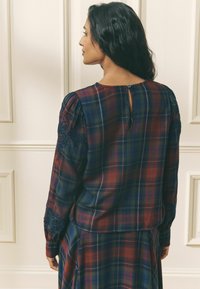 Rutigt blus med mörkblå, röd och grön färg, puffärmar, blommig broderi på axeln och en nyckelhålsstängning på baksidan.