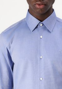 Lyseblå knapp-up skjorte laget av teksturert stoff, med klassisk krage, hvite knapper og en liten brodert logo på brystet.