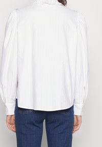 Camicia bianca con maniche a sbuffo e colletto con pizzo, caratterizzata da un taglio dritto e chiusure a bottoni nei polsini, abbinata a denim scuro.
