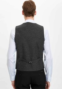 Uomo con un gilet formale grigio scuro indossato sopra una camicia formale azzurro chiaro, abbinato a pantaloni neri, visto da dietro.