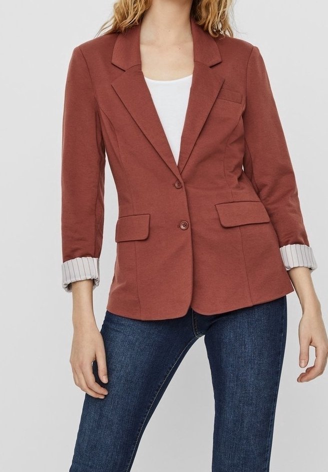 Femme portant un blazer rouille boutonné avec des poignets rayés retroussés, un haut blanc et un jean bleu foncé sur un fond uni.