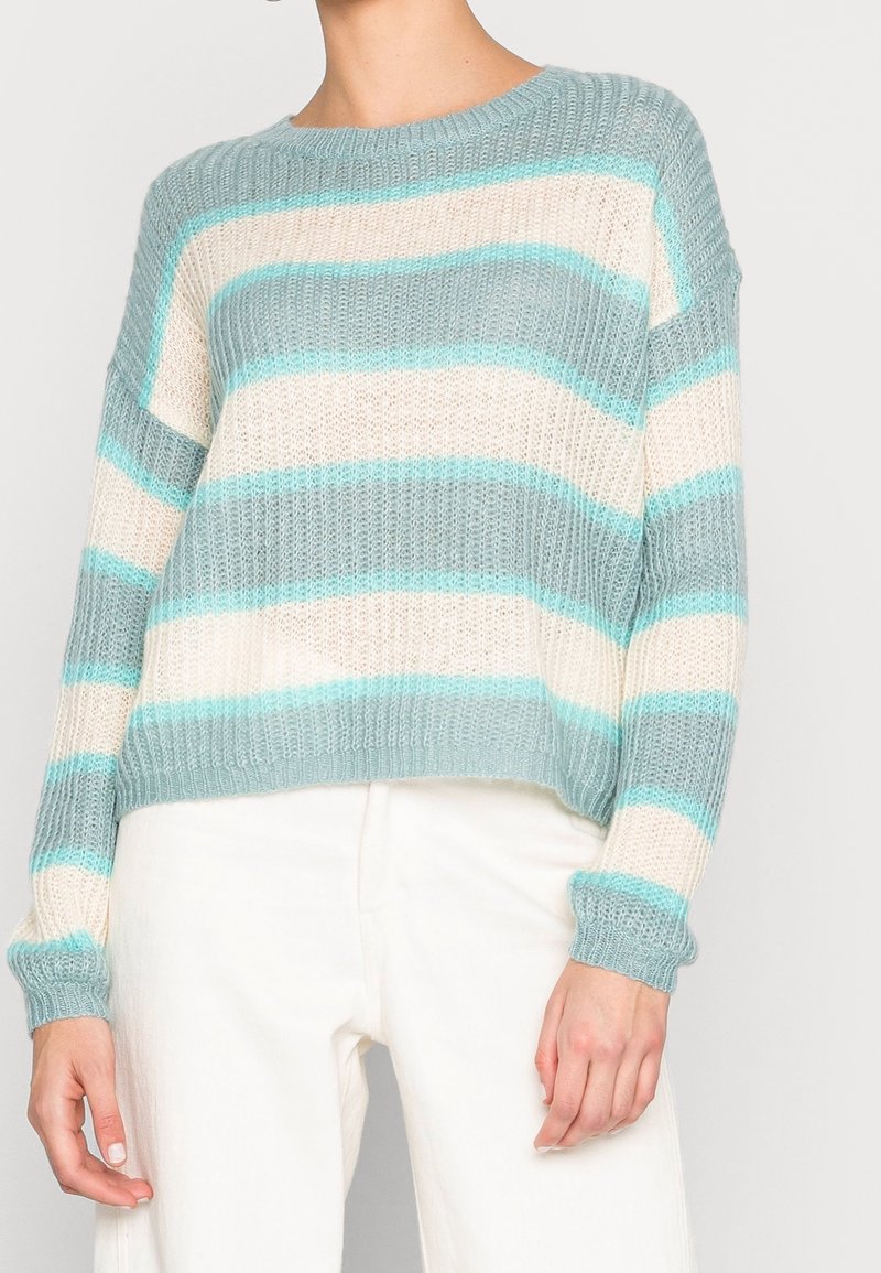 Personne portant un pull en maille lâche à rayures horizontales turquoise et crème, associé à un pantalon blanc taille haute, sur un fond uni.