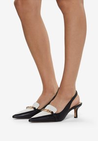 Black and white slingback heels with pointed toes, featuring a gold chain accent and a low heel. Smooth leather texture and contrasting color blocks.